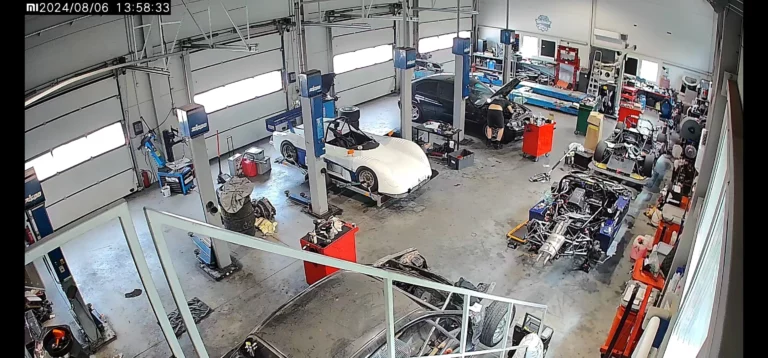 Warsztat samochodowy z kilkoma samochodami na podnośnikach, w tym białym samochodem sportowym i czarnym samochodem z otwartą maską. Widać również podwozie samochodu wyścigowego. Narzędzia i sprzęt są rozłożone po całym warsztacie.