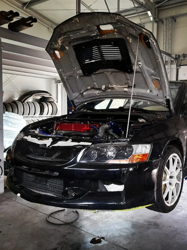 Otwarte maska czarnego samochodu sportowego, ukazująca czerwony pokrywa silnika i liczne komponenty w komorze silnika, na tle warsztatu z oponami i sprzętem motoryzacyjnym.