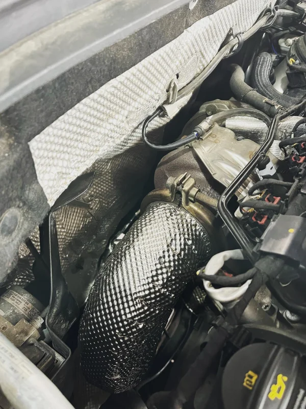 Zbliżenie na układ wydechowy w komorze silnika, z widoczną owijką termiczną na rurze wydechowej oraz otaczającymi komponentami silnika i osłoną termiczną.