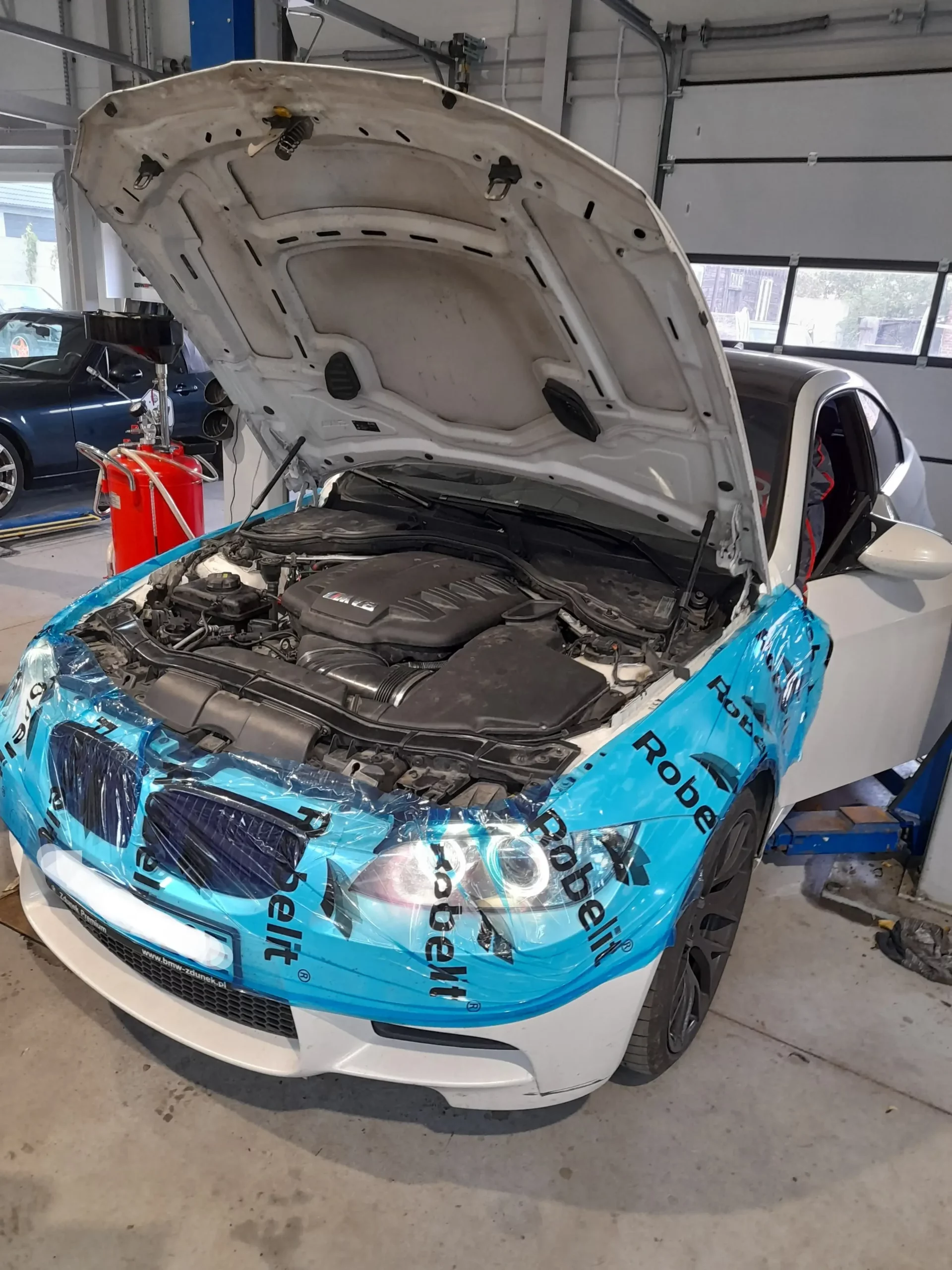 Na zdjęciu widzimy białe auto, umieszczone na podnośniku w warsztacie samochodowym. Samochód ma otwartą maskę, co umożliwia dostęp do silnika, który jest widoczny. Przednia część pojazdu jest pokryta niebieską folią ochronną z napisem "Robelit", chroniącą lakier przed uszkodzeniami podczas pracy. W tle widoczne są różne narzędzia i sprzęt warsztatowy, w tym czerwona skrzynka narzędziowa oraz inne pojazdy, co sugeruje, że warsztat jest dobrze wyposażony i aktywnie wykorzystywany do napraw i konserwacji pojazdów. Atmosfera zdjęcia wskazuje, że pojazd jest w trakcie przeglądu lub naprawy technicznej.