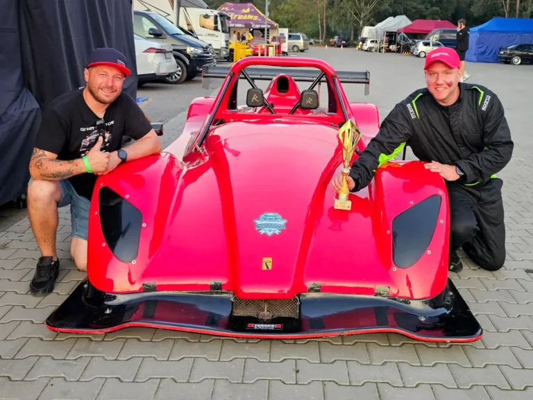 Dwóch mężczyzn pozuje przy czerwonym samochodzie wyścigowym. Jeden z nich, w czarnym t-shircie i szortach, stoi obok samochodu, a drugi, ubrany w kombinezon wyścigowy, trzyma trofeum. W tle widoczne są inne pojazdy oraz namioty, co sugeruje udział w zawodach wyścigowych.