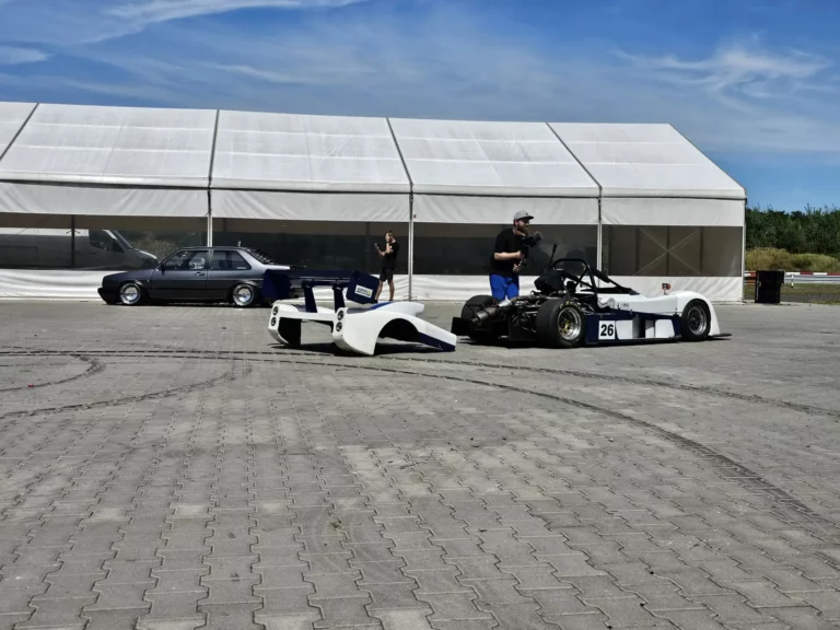 Widok na prawy tył samochodu wyścigowego Radical SR3 z numerem 26, ukazujący szczegóły zawieszenia, układu hamulcowego oraz opony. W tle widoczny jest biały namiot oraz narzędzia warsztatowe.