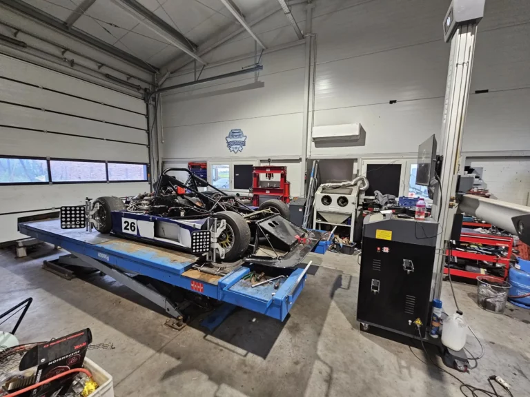 Widok na Radical SR3 z numerem 26, umieszczonego na podnośniku w warsztacie. Ukazany jest szczegół zawieszenia, układu hamulcowego oraz opony Pirelli. W tle znajduje się dobrze wyposażony warsztat z narzędziami i sprzętem.