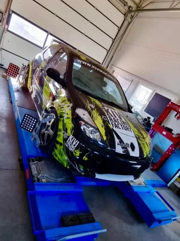 Samochód wyścigowy w unikalnym czarno-żółtym malowaniu z napisem "KIWI RACING FACTORY" podczas testów na niebieskiej platformie w warsztacie MC Garage. Widać podłączone do kół urządzenia oraz sprzęt w tle, co sugeruje testy wydajności lub wyrównania.